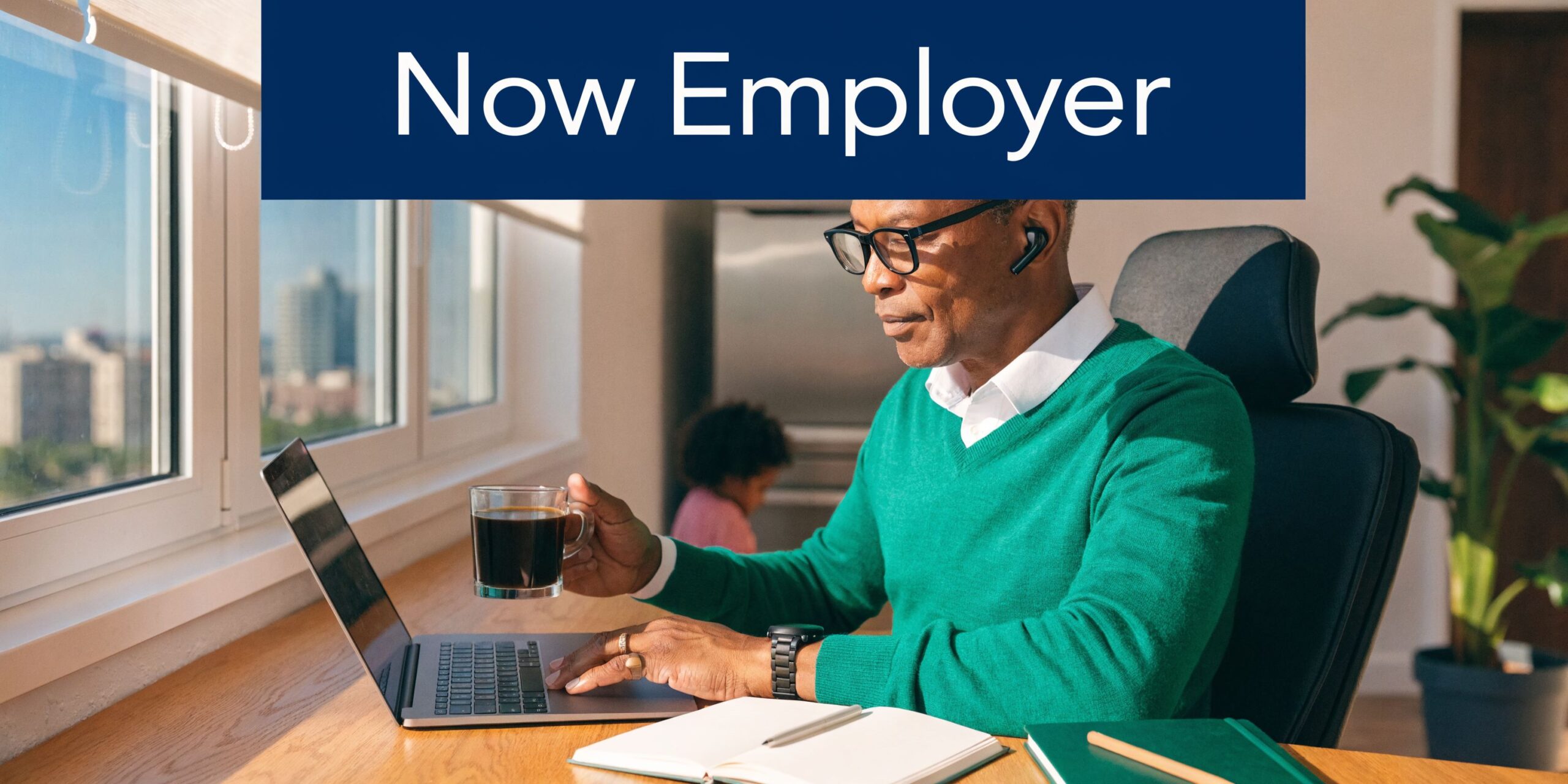 A professional man wearing glasses and a green sweater working on his laptop while drinking coffee at home.