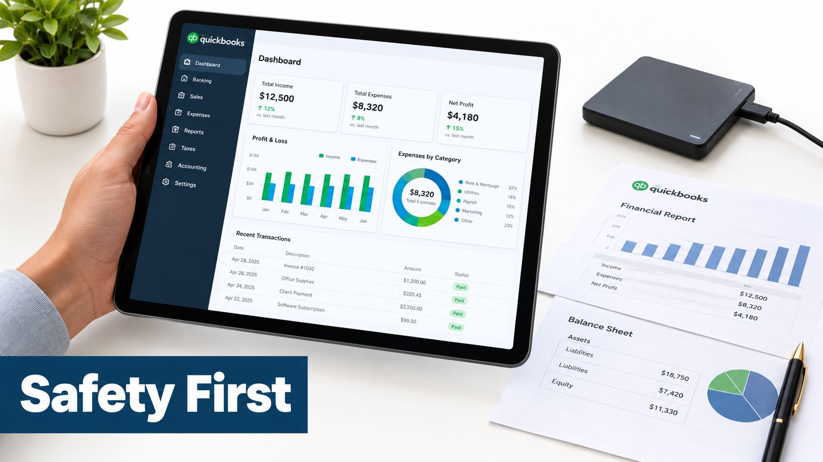 A hand holds a tablet displaying a QuickBooks financial dashboard beside paper reports and a hard drive.